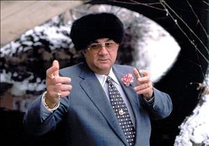 Marks wearing a suit, multiple rings, corsage, tie pin, bracelet, watch, tinted sunglasses and black Ushanka, gesturing dramatically towards the camera