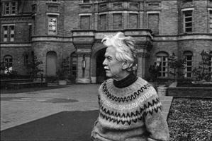 An older Morgan posing in profile in front of a large, landscaped brick building, wearing a circular patterned sweater