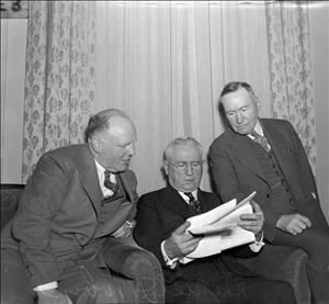 Beck leaning over a seated man holding loose sheets of paper as another man sits on the arm of the chair to read along