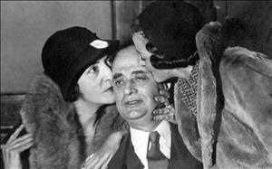 Pantages being kissed on either side of the head by two women wearing fur coats and hats making eye contact with one another