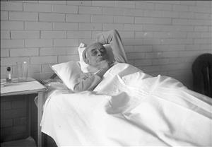 Pantages resting his hand on his chin, laying in a bed under a white sheet beside a coffee table with a medicine jar and an empty glass in front of a bare brick wall