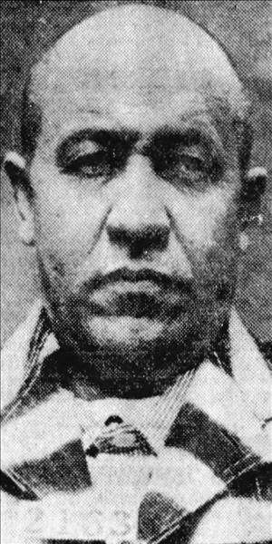 Hillman wearing a striped prison uniform with closely cropped balding hair