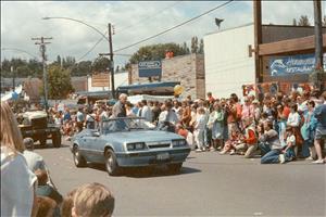 Woodward standing inside a small, blue convertible sports car in front of parade attendees on a street with street signs reading Winslow Hardware, Paper Products, etc. and Hemingway Restaurant
