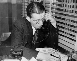 Woodward on the phone with slicked back hair inspecting a newspaper with a pencil in his hand