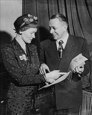Walter and Mildred Woodward smiling broadly, looking at a newspaper page in front of a long curtain