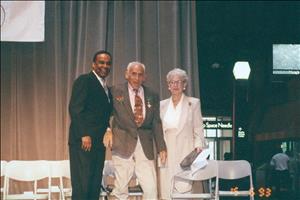 Hans and Thelma Lehmann standing with a smiling man on a stage in front of a row of folding chairs, an exit that reads Space Needle in the background
