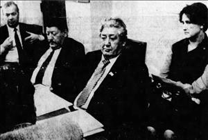 Lavroff in a suit and tie with eyes cast downward while sitting at a desk with two other people and one person in the background
