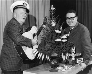 McCune smiling in thick rim glasses leaning on a desk decorated with a tree strung with fish ornaments, a faux-Native American sculpture holding a sign reading "Keep Clam" next to a man in a Navy har with an acoustic guitar in his hands