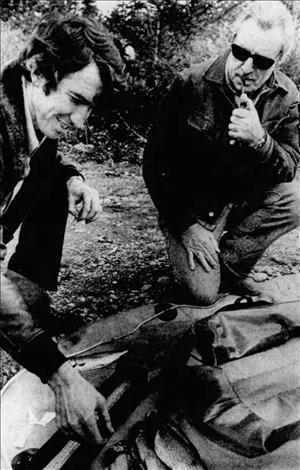 An older McCune wearing sunglasses holding a pipe, kneeling next to a man reaching for fish