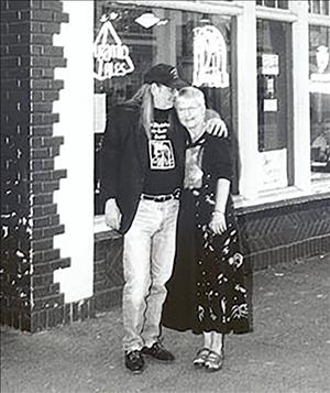 A man in a black baseball cap with his arm around a woman with short hair in front of a storefront with ornate brick patterns and neon signs in the window