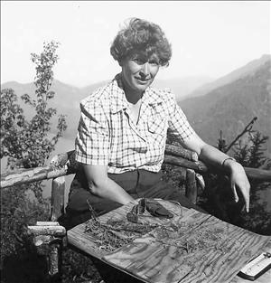 Buxbaum sitting outside on a patio with a rough hewn wooden guardrail in a button down shirt in front of a forested, mountainous setting