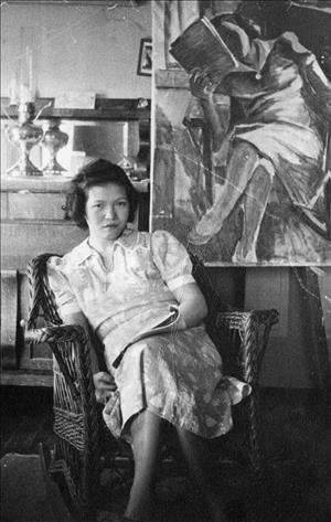 Bernadette Horiuchi posing in a wicker chair wearing a dress with a magazine folded in her hand in front of a painting of her by Paul Horiuchi in a similar pose with an ornate brass lantern in the background