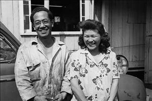 Paul Horiuchi posing in a dirty jumpsuit beside Bernadette in a floral dress in front of a vehicle 