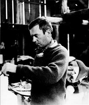 McCracken in a barn with gaps in the roof and panelling painting with a brush and wearing a sweater