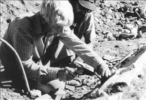 Wehr crouched in a plaid shirt with white hair, next to a man with a beard wearing sunglasses and a baseball cap, chiseling at a slab of rock