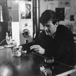 Weir looking at a white coffee cup that he has balanced a pair of glasses on the handle as he sits at a table beside an ashtray and a pack of cigarettes