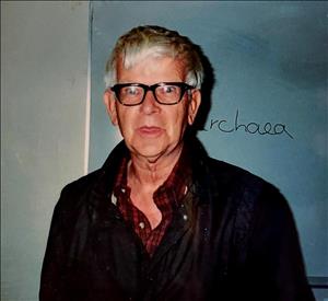 Weir posing in front of a wall with hand writing on it, wearing a plaid shirt and thick rim glasses