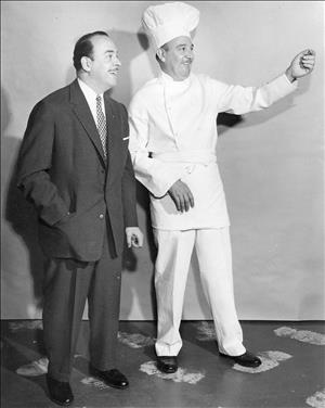 Rosellini smiling with his hand tucked into his suit pocket beside a man in chef's whites