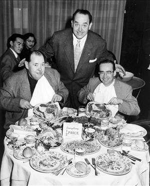 Rosellini smiling in a three piece suit over two seated diners with white napkins tucked into their collars and cutlery raised to their plates behind a full table of food and a sign reading "Everything 1/2 Price"
