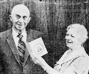 Helfgott smiling in front of a wood paneled wall while holding a small booklet up beside to a man in a pinstripe suit and tie