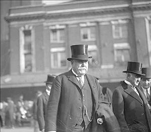 Hill in a three piece suit and top hat walking outside among similarly dressed men