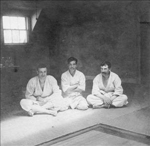 Three men sitting with crossed legs in an unlit room with one window wearing white judogi uniforms