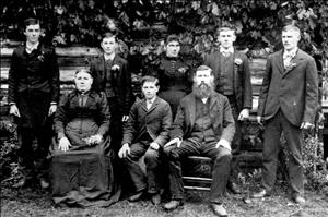 A family of eight posing outside, in front of a log cabin wearing dark formal wear