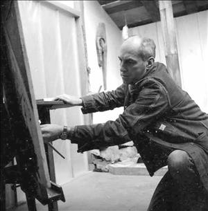 Anderson in a denim jacket kneeling with a paint brush against canvas