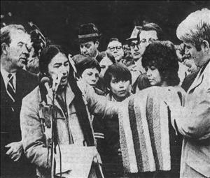 Whitebear speaking into multiple microphones as he hands a striped blanket to a man in a suit as children, a woman and journalists watch in the background