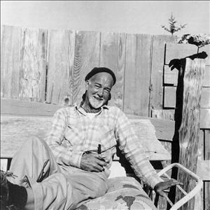 Callahan smiling in a knit cap and plaid shirt holding onto a pipe and reclining on patio furniture