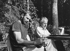 Graves sitting with another man outside at a table, in front of a natural wooded landscape