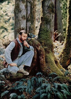 Graves posing in a vest, collar shirt and sneakers, kneeling with his hand on his chin in the middle of a mossy forest