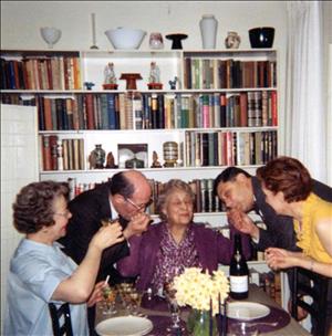Dusanne in a floral patterned shirt and purple sweater being kissed on both hands by two men in suits as two other women smile and clap inside a dining room lined with books and ceramic art