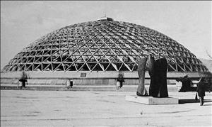 A triodetic dome the size of a small arena in front of a rectangular pool filled with lily pads and conifer trees on either side of the structure