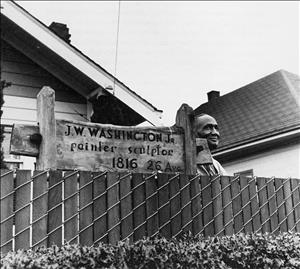 Washington smiling behind a fence, beside a sign reading "J.W. Washington Jr., painter, sculptor, 1816 26 Avenue"