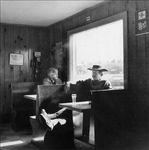 Robbins in cowboy hat and poncho leaning in a booth with a glass of beer during the day