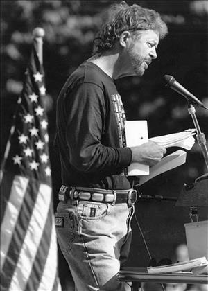 Robbins with a short beard holding papers and speaking into a microphone with an American flag in the background
