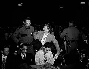 Florence James being pulled up aggressively by a police officer in a theater while members of the row in front of them bow their heads