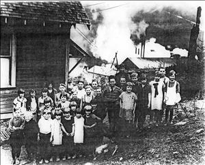 Group of school children outside of a small wooden building in front of massive plumes of smoke in the background