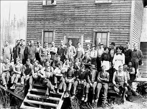 Group of mainly men and two women in work clothes, sitting and standing on a porch in front of a wood paneled building