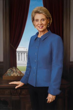 A painting of Gregoire in a blue button down shirt and black pants with a hand on a wooden desk in front of an open window where we can see a white, columned government building in the background