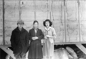 An elderly Japanese man smoking a cigarette next to an elderly Japanese woman both in winter clothing standing next to a younger Japanese woman in a white dress, in front of a wooden wall 