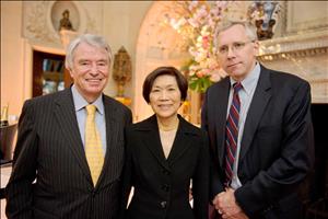An older Bonker smiling in yellow tie and pinstripe suit jacket posing with two other people inside of an ornately decorated building with champagne and flowers in the background