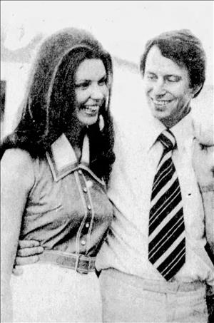 Bonker with jacket over his shoulder smiling with his wife outside, wearing a sleeveless shirt with a broad collar