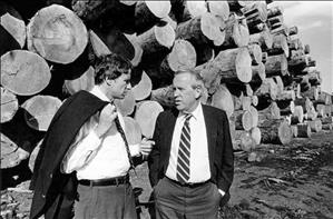 Bonker with jacket over his shoulder speaking with another man in front of a wall of stripped timbre stacked about ten trees high