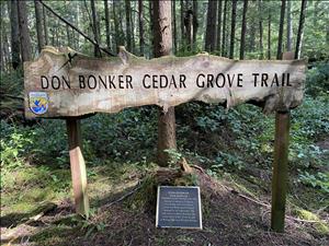 A sign made from a slice of timber reading "Don Bonker Cedar Grove Trail" above a small bronze and black plaque in front of a wooded area