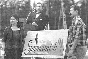 Two people pose in front of a sign that reads "City of Sammamish, Washington" in front of a person behind a podium speaking into a microphone