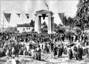A crowd gathered around a rectangular stone archway with hanging pendants and flags and houses in the distance 