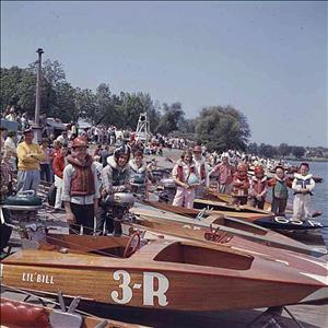 Young adults, children and adults stand on the shoreline beside boats wearing life preservers, baseball and football helmets, with a boat in the foreground with the words "Lil' Bill 3-R" painted on the side