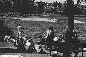A crowd in a park setting watches motorized boats moving quickly on the water in the background